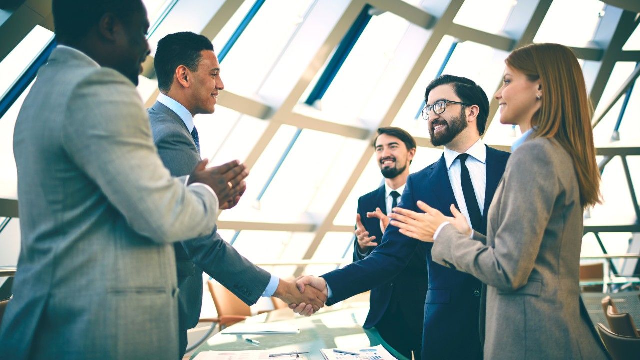 Executives shaking hands in a daylit glass-roofed meeting space.