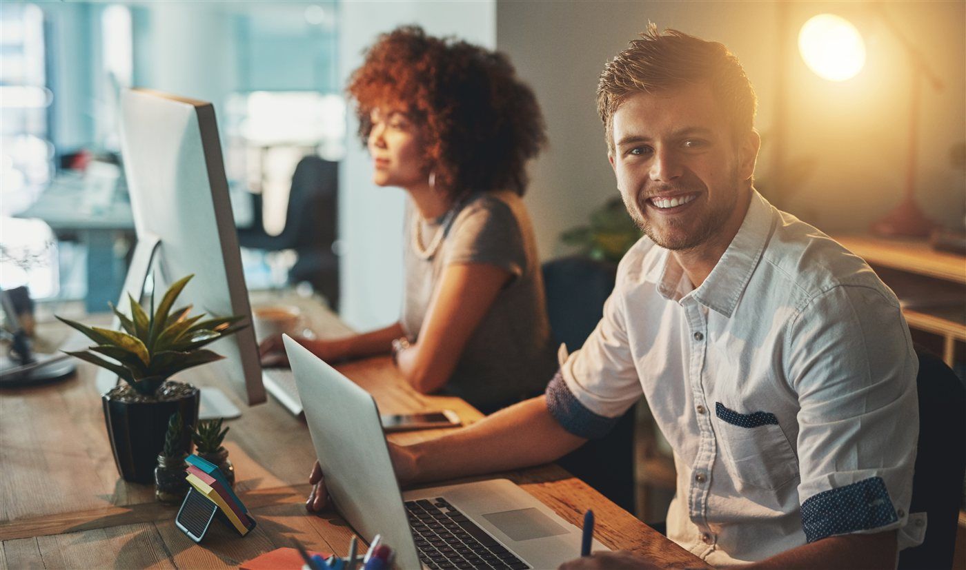 A smiling man and a colleague working at desks in warm evening light.