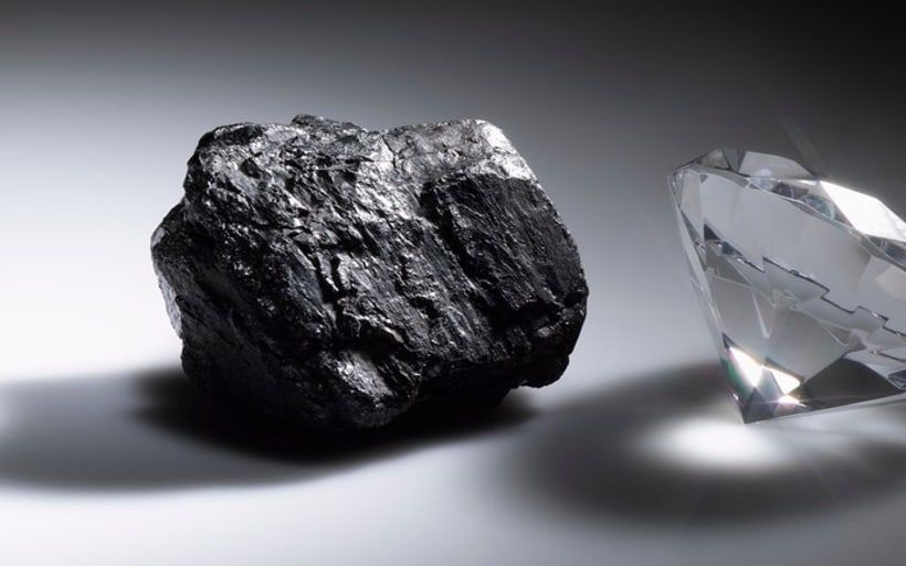 A rough lump of coal lit against a dark ground, a cut diamond just beyond it.