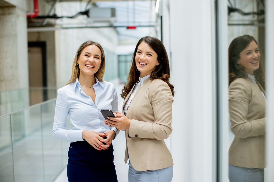 Two professionals talking beside a glass partition in a bright office corridor.